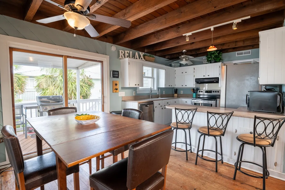 A bright, airy dining area with a view toward the outdoors, featuring a table with bar stools, modern kitchen cabinetry, and large windows that allow natural light to fill the space. The scene suggests easy access to the deck or porch, inviting guests (and their pups) to flow between indoor and outdoor living.