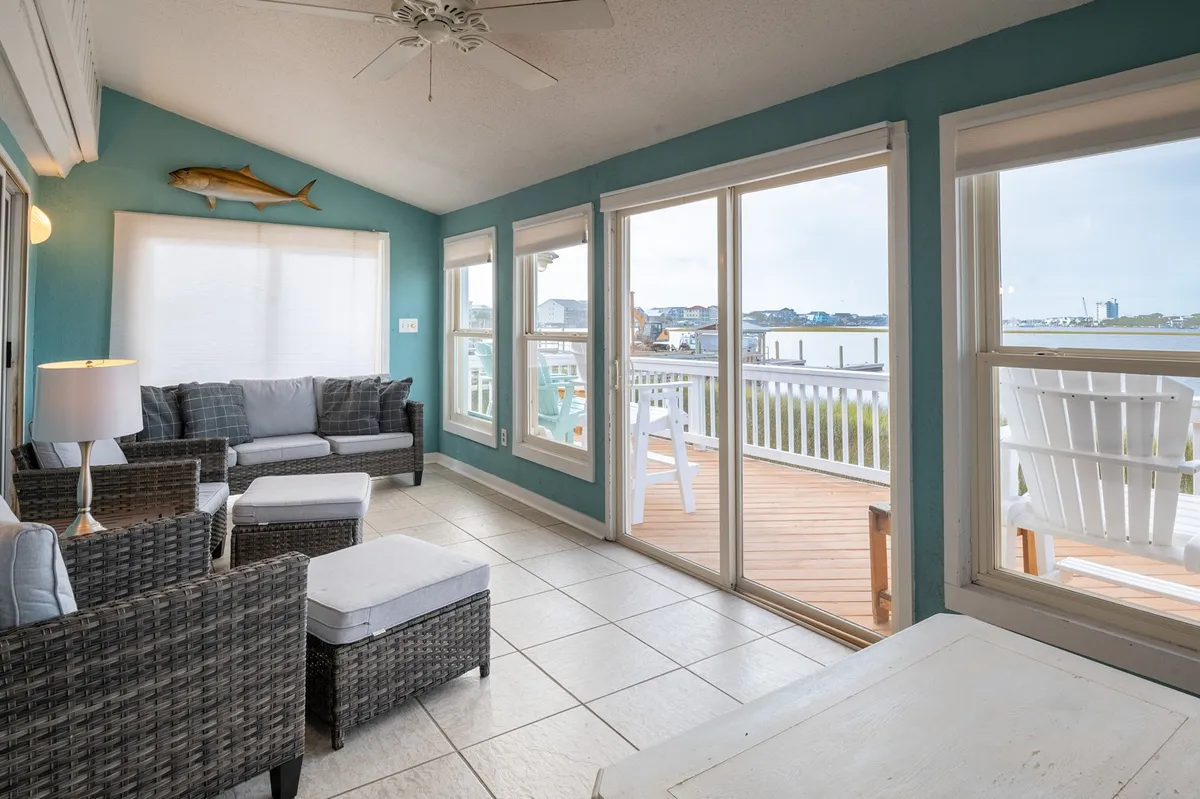 A bright Florida room with wicker furniture, large panoramic windows that offer sweeping water views, and coastal-inspired fishing décor. Light-colored walls and comfortable seating create a serene lounge atmosphere.