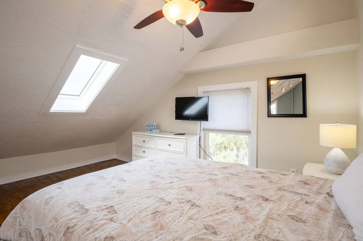 A cozy loft bedroom with a bed topped by patterned bedding, a ceiling fan above, a TV atop a dresser, and a skylight that lets natural light flood the space. The lamp and soft accents further enhance the restful appeal.