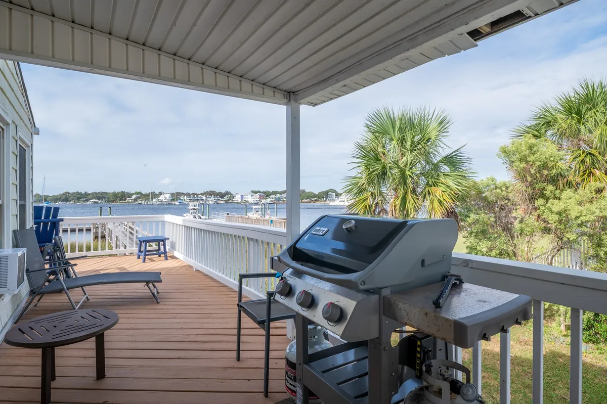 An inviting deck area features a gas grill and a comfortable seating arrangement, all overlooking a stunning water view with palm trees for shade. The setting is ideal for waterfront barbecues, morning coffee, or evening gatherings with friends and family.