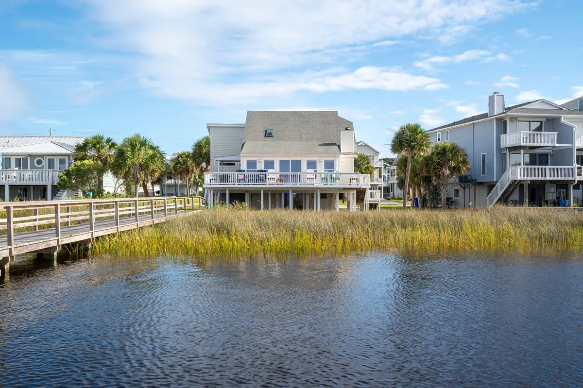 A waterfront house with a wooden boardwalk running through a lush, grassy yard and surrounded by palm trees. The scene captures the proximity to the water, the inviting boardwalk leading to the dock, and the spacious outdoor area perfect for parking and prepping gear.