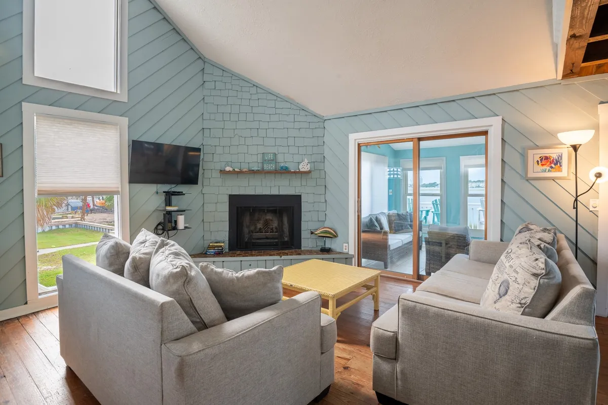 A cozy, sunlit living room with plush sofa seating arranged around a decorative fireplace. Large windows invite in natural light, and curated decorative items on the shelf create a welcoming and homey atmosphere.
