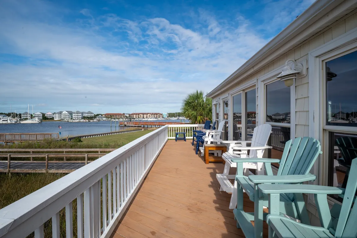 A spacious wooden deck wraps around the waterfront side of the home, furnished with Adirondack chairs that face panoramic water views. Palm trees frame the setting, and the tranquil expanse of water stretches to the horizon, offering a perfect vantage for enjoying sunrises, sunsets, and gentle boat traffic.