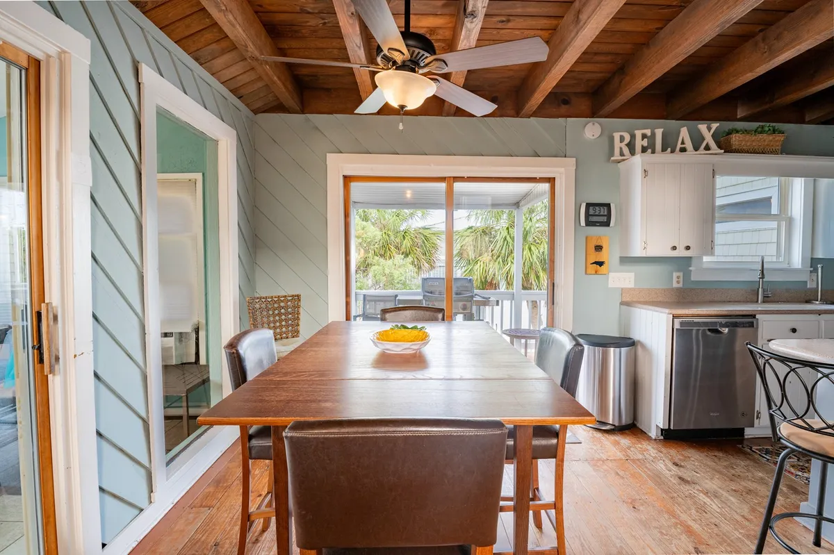 A sunny, inviting dining area with a large table set with a fruit bowl, ceiling fan above, and expansive windows framing lush greenery. The atmosphere is relaxed and casual, perfect for family meals or winding down after a day exploring the beach and boardwalk.