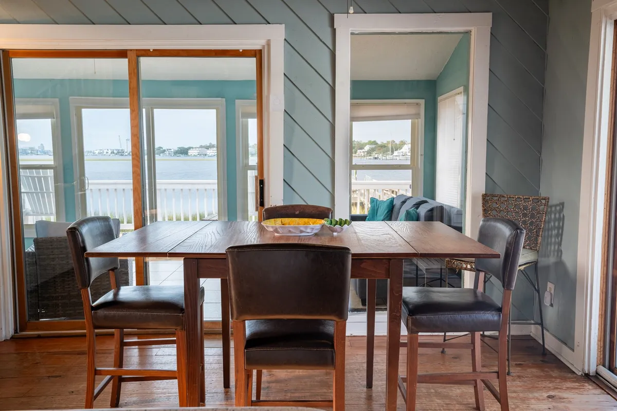 A wooden dining table surrounded by chairs sits in front of large windows that showcase scenic water views. A decorative bowl of fruit adds a cheerful touch, enhancing the room’s inviting and communal feel.