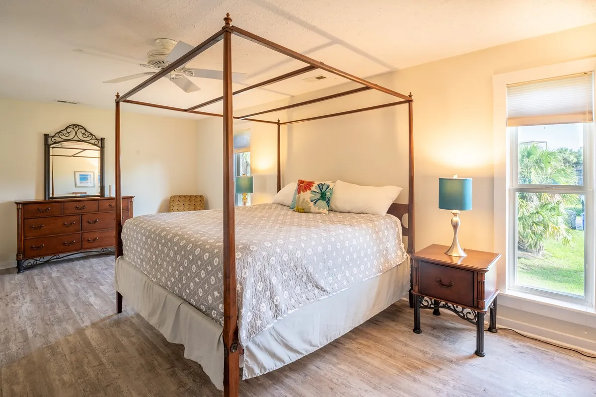A spacious bedroom featuring a four-poster bed dressed with plush decorative pillows, a nightstand with a warm lamp, and a large dressing mirror. Soft sunlight filters through the windows, filling the space with a tranquil and intimate atmosphere.