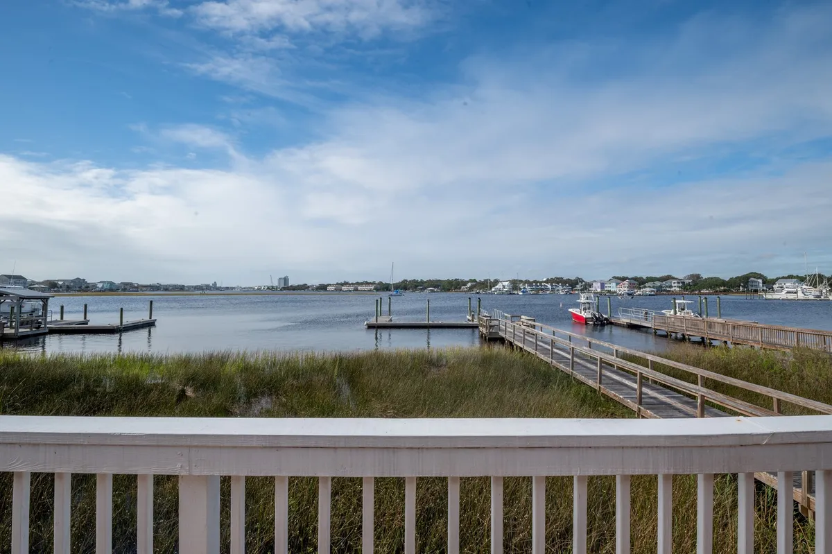 A serene perspective of calm water, docks, and lush marsh grass under a wide blue sky. The vantage reveals anchored sailboats and gentle boat activity, encapsulating the peaceful sights and sounds that define waterfront living.