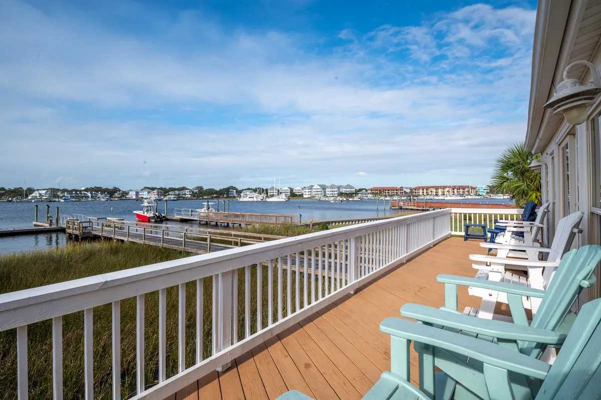 The image features an inviting waterfront porch overlooking tranquil marina waters. Adirondack chairs line the porch, offering picturesque views of anchored sailboats and a peaceful boardwalk. The scene is serene, with gentle sunlight illuminating the outdoor seating, perfect for relaxing with coffee or watching passing boats.